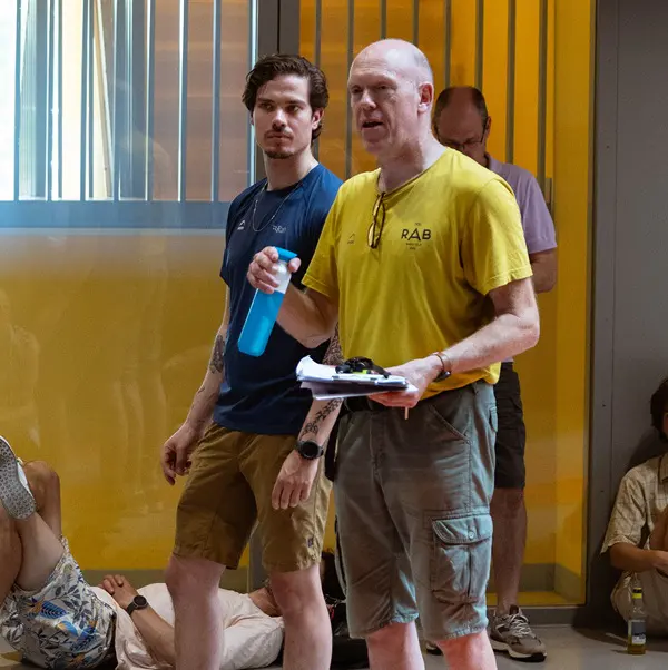 Twee mannen staan binnen; de ene houdt een blauwe waterfles vast en de andere heeft papieren en een zonnebril in zijn handen. Op de achtergrond zitten of liggen mensen op de vloer. De ruimte heeft gele muren en glazen panelen.